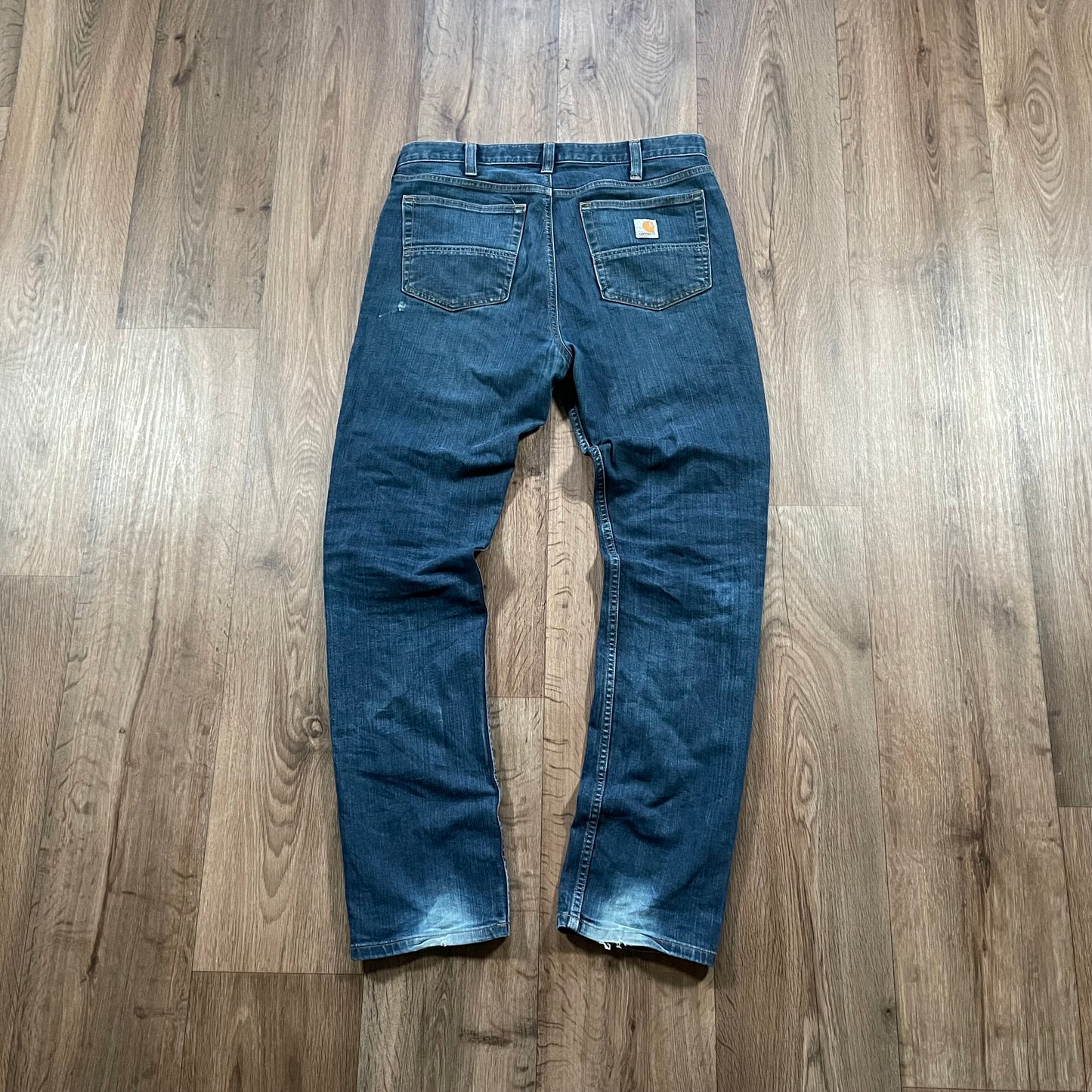 A pair of Madams Vintages Carhartt Dark Wash Jeans is laid flat on a wooden floor, featuring a small brown leather patch on one back pocket, subtle fading on the legs and hems, and distressed detailing that adds to their rugged charm.