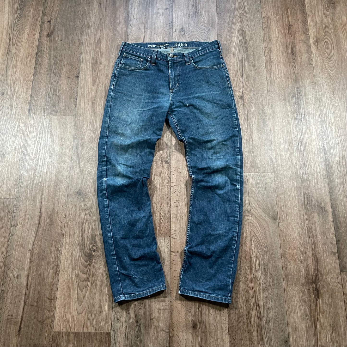 A pair of Carhartt Dark Wash Jeans by Madams Vintage lies flat on a wooden floor, showcasing distressed detailing that enhances natural creases and color fading, particularly around the knee and thigh areas.