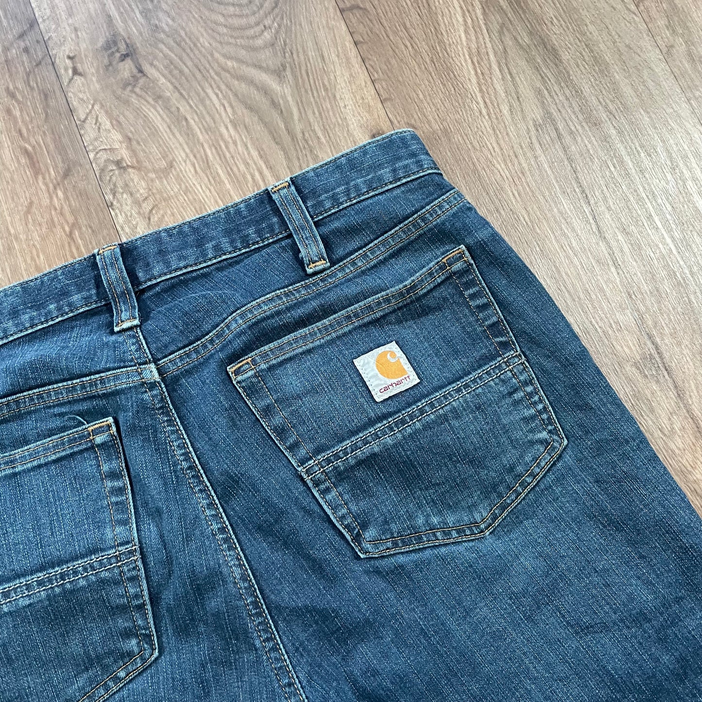 Carhartt Dark Wash Jeans by Madams Vintage are lying on a wooden floor, featuring a Carhartt logo patch on the back pocket, visible stitching, belt loops, and subtle distressed detailing for added character.