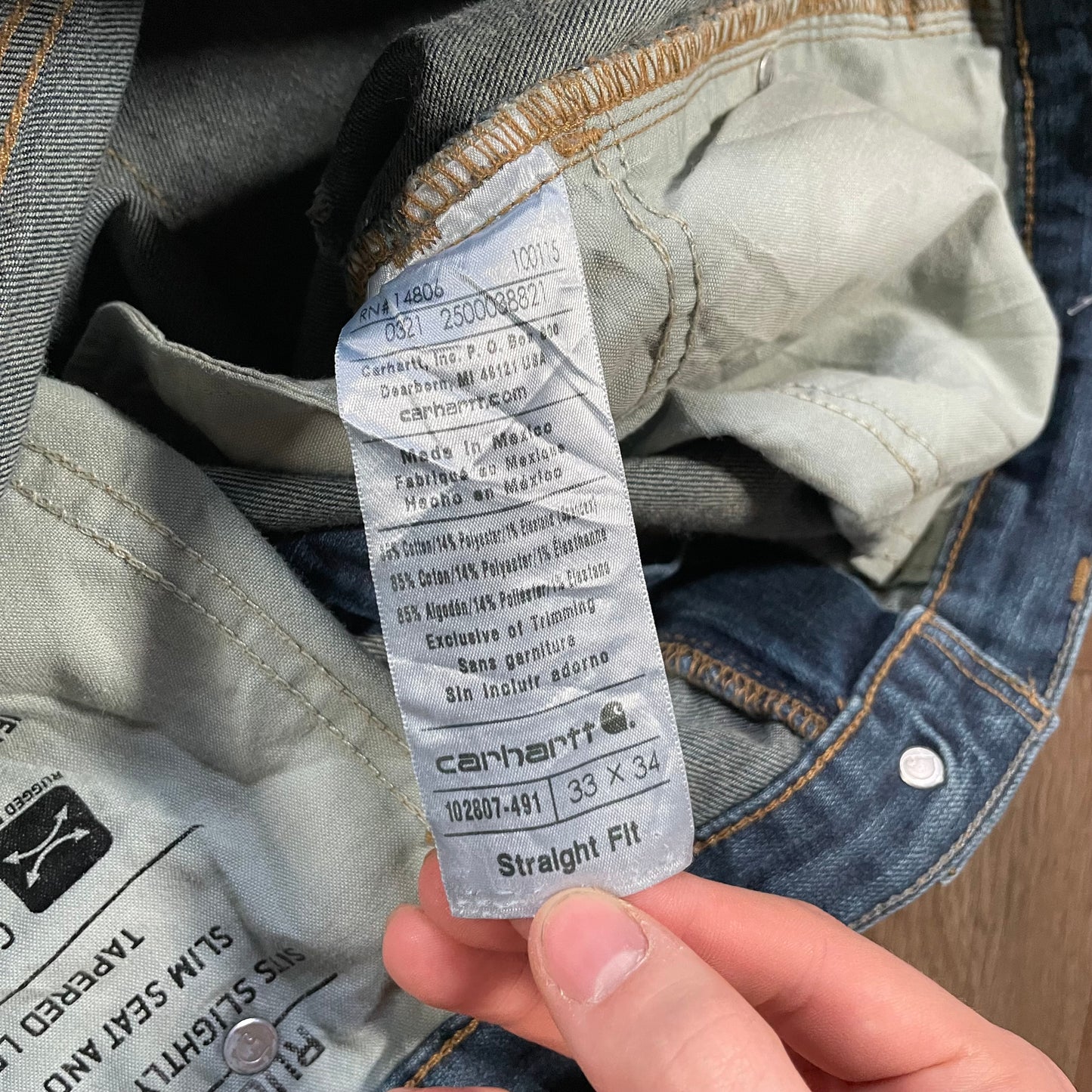 A person holds a care tag from Madams Vintages Carhartt Dark Wash Jeans, displaying the washing instructions, material composition, size 33 x 34, and Straight Fit style. The blue jeans feature visible stitching.