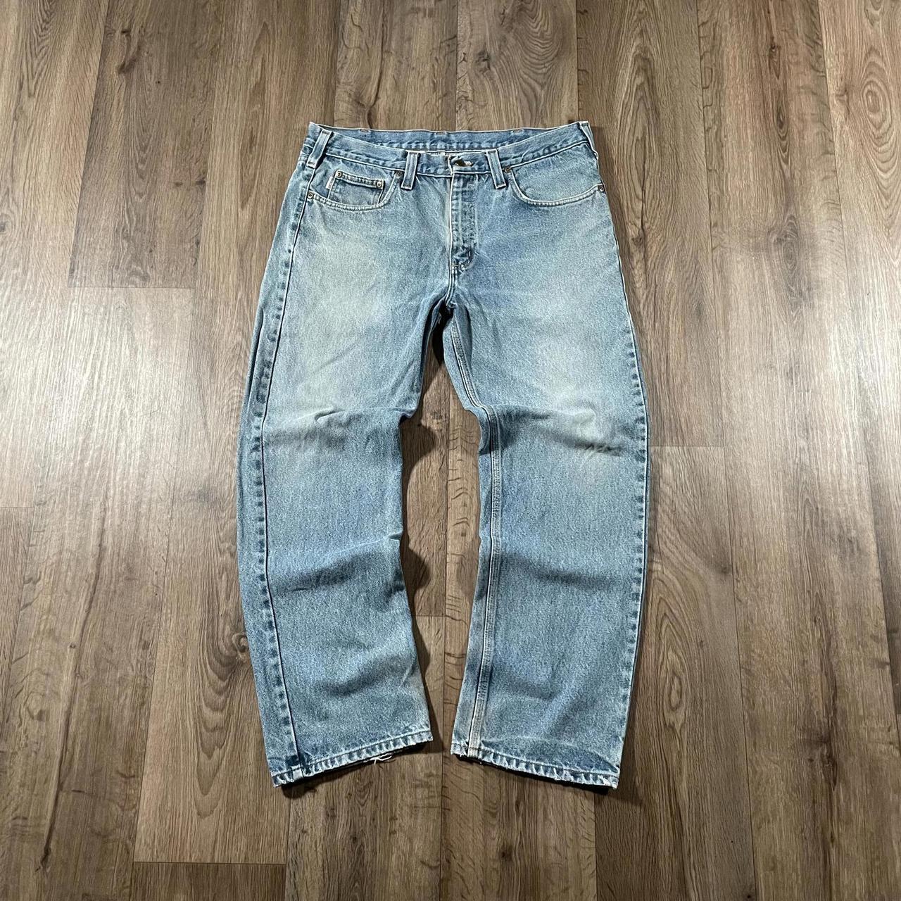 The Carhartt Blue Denim Jeans W36 L30 by Madams Vintage are laid flat on a wooden floor. Their classic five-pocket design, visible stitching, and slightly worn look contribute to an authentic workwear vibe, while creasing at the knees and ankles enhances their timeless appeal.