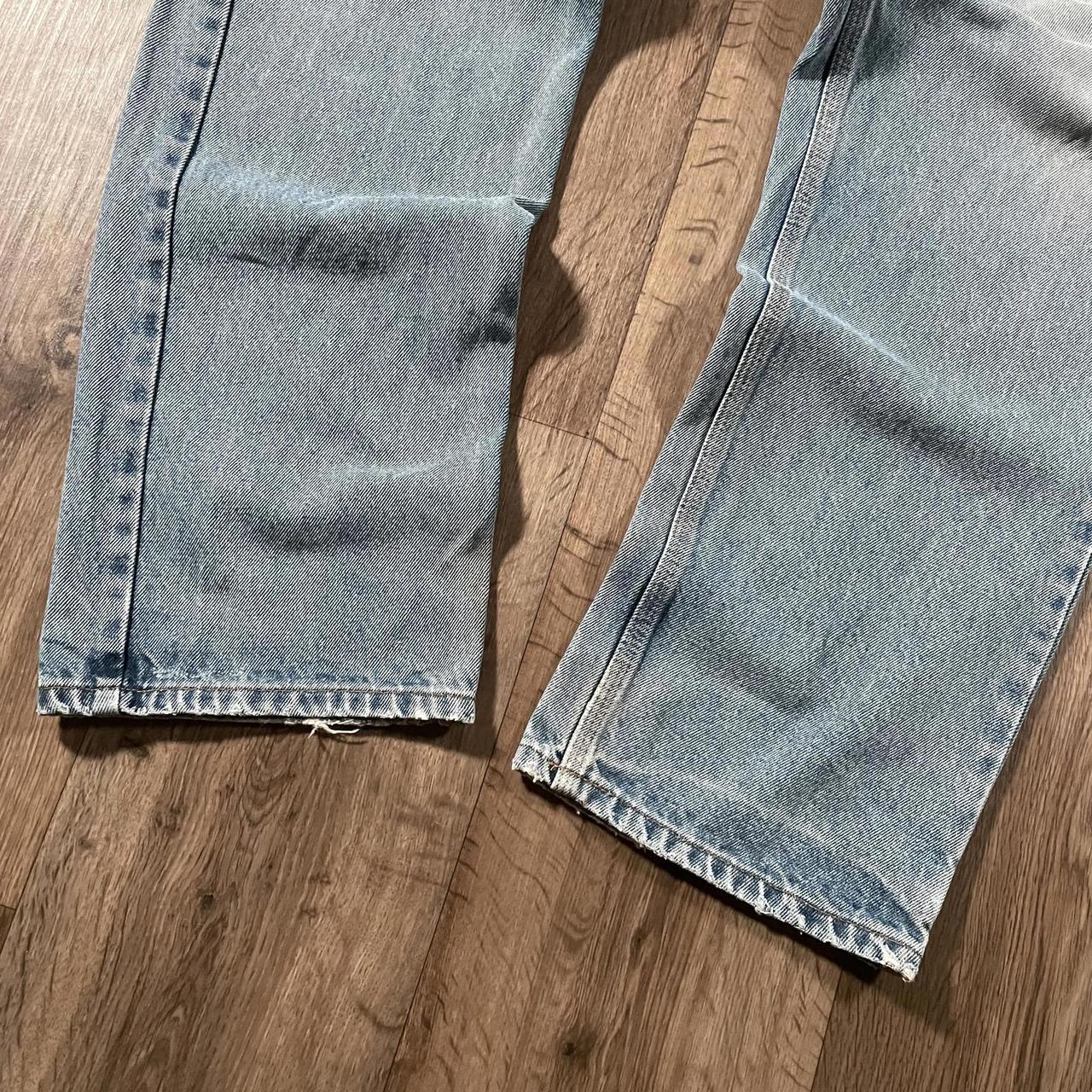 Close-up of Madams Vintage Carhartt Blue Denim Jeans W36 L30 on a wooden floor, featuring slightly faded legs, natural creases, and visible fringed hems.
