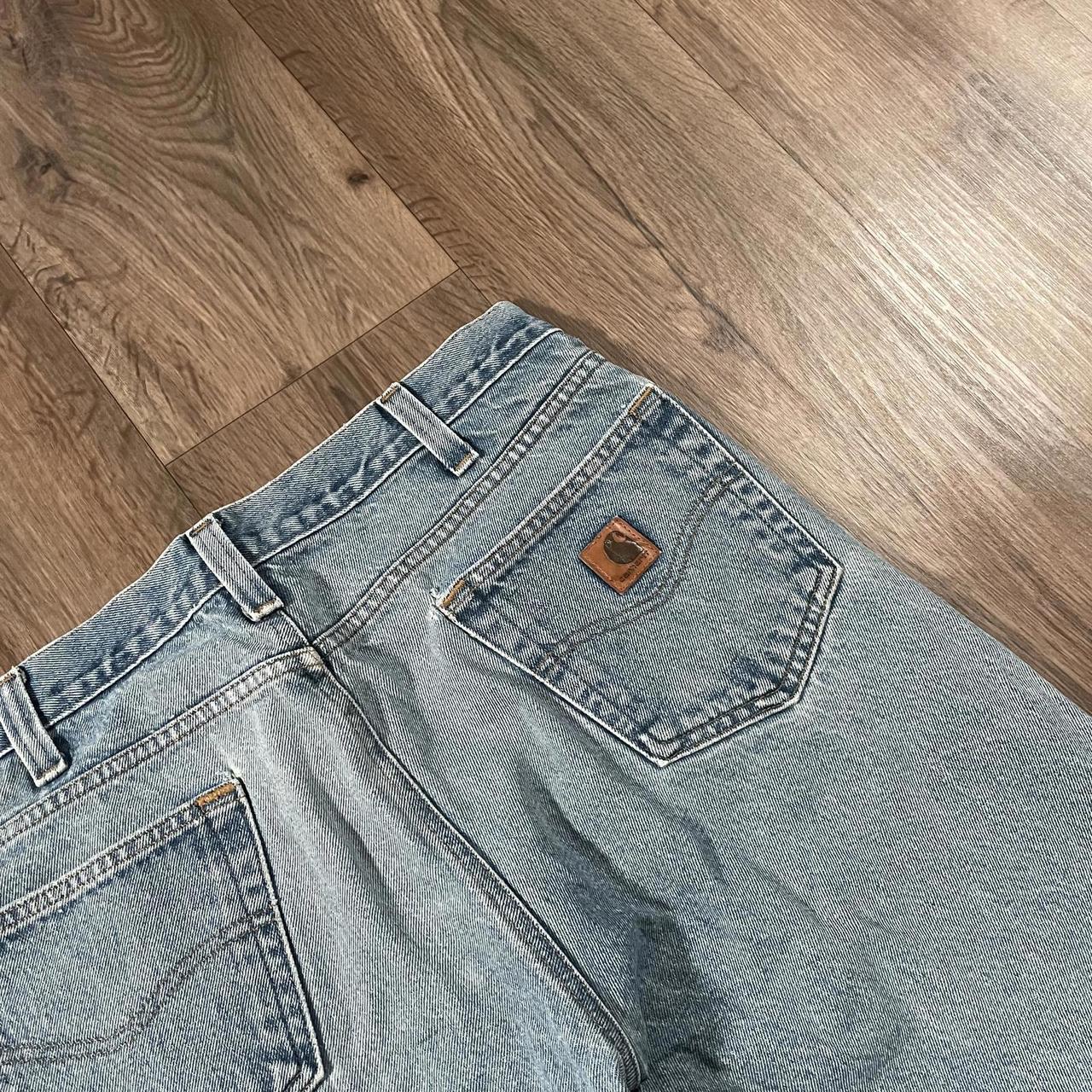 Lying on a wooden floor, the Carhartt Blue Denim Jeans W36 L30 from Madams Vintage showcase their back side with a brown patch above the pocket, elegantly highlighting the pocket and belt loops and embodying classic workwear style.