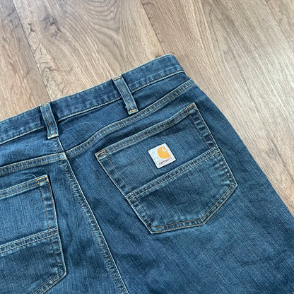 Carhartt Dark Wash Jeans by Madams Vintage are lying on a wooden floor, featuring a Carhartt logo patch on the back pocket, visible stitching, belt loops, and subtle distressed detailing for added character.