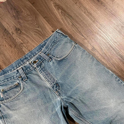 The Madams Vintage Carhartt Blue Denim Jeans W36 L30 rest on a wooden floor. They showcase a classic five-pocket design with visible stitching and a button closure, capturing timeless workwear style.