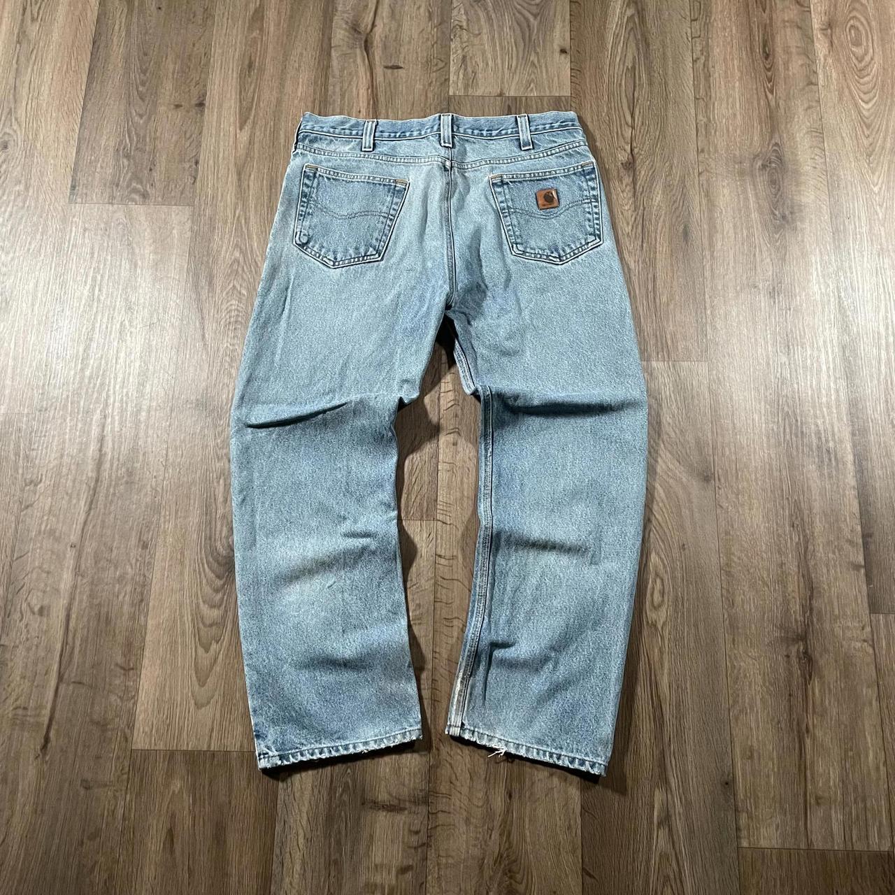 The Madams Vintage Carhartt Blue Denim Jeans W36 L30 are laid flat on a wooden floor. These jeans have light blue color with a leather patch on the back pocket, visible stitching, and a slightly faded look at the knees, showcasing their classic workwear style.