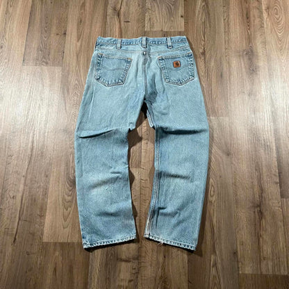 The Madams Vintage Carhartt Blue Denim Jeans W36 L30 are laid flat on a wooden floor. These jeans have light blue color with a leather patch on the back pocket, visible stitching, and a slightly faded look at the knees, showcasing their classic workwear style.