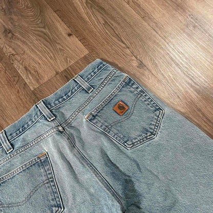 Lying on a wooden floor, the Carhartt Blue Denim Jeans W36 L30 from Madams Vintage showcase their back side with a brown patch above the pocket, elegantly highlighting the pocket and belt loops and embodying classic workwear style.